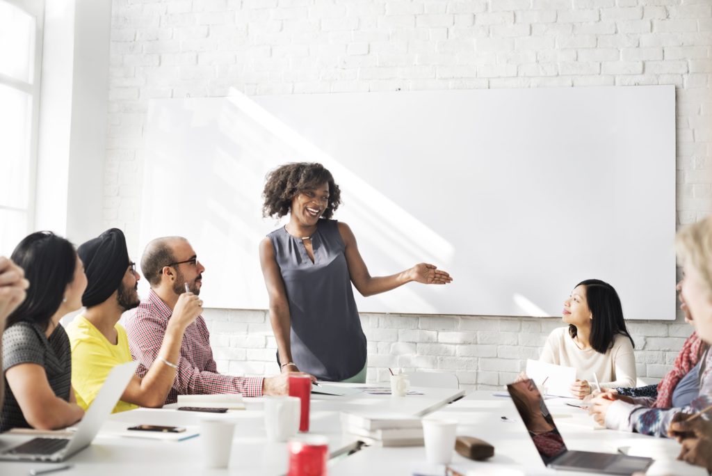 A woman stands in front of a meeting of people, addressing them.