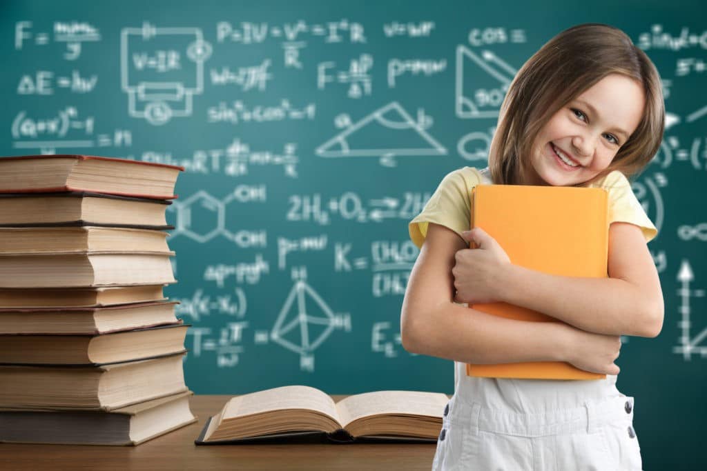 Young girl smiling and clutching a book in front of a desk and chalkboard.
