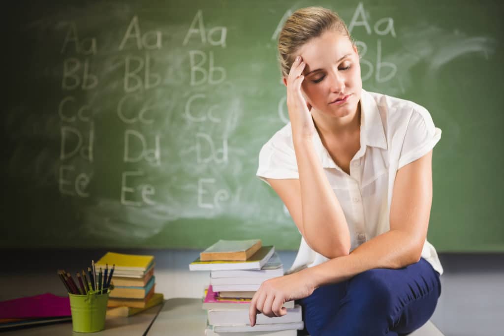 Frustrated teacher sitting on a desk in front of a chalkboard.