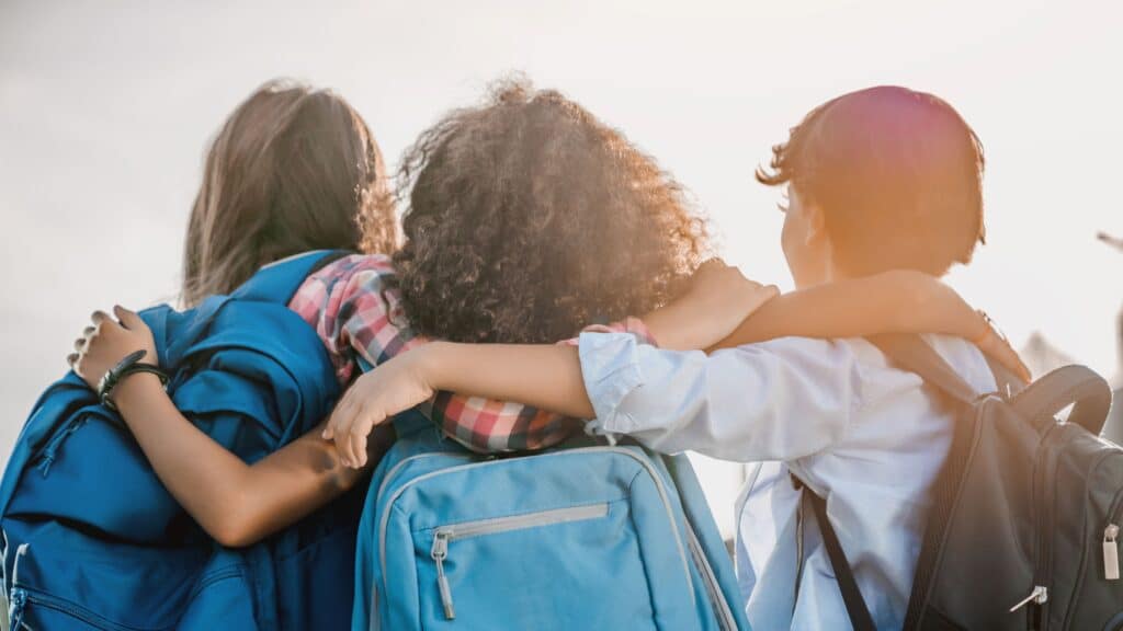 Rearview shot of three students standing with their arms around each other, wearing school backpacks.