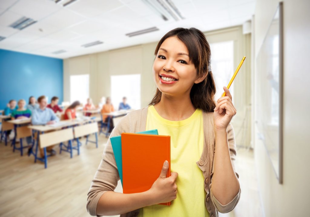 A female teacher stands smiling with her book and pencil in front of her classroom.