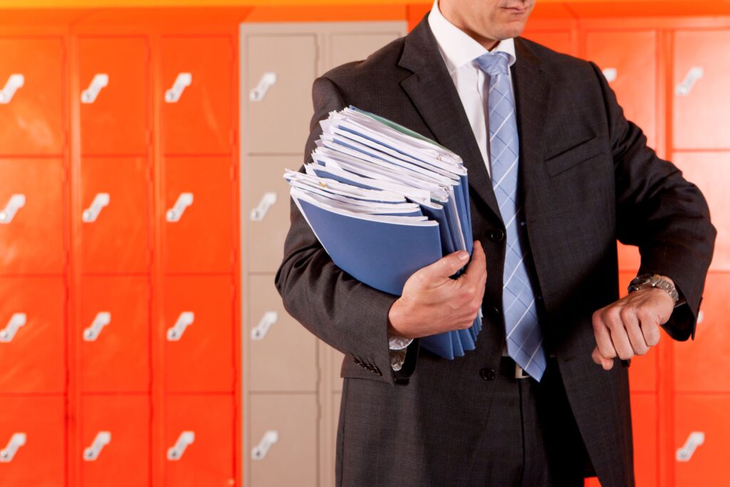 An educator holds a stack of folders and checks his watch in the hallway.