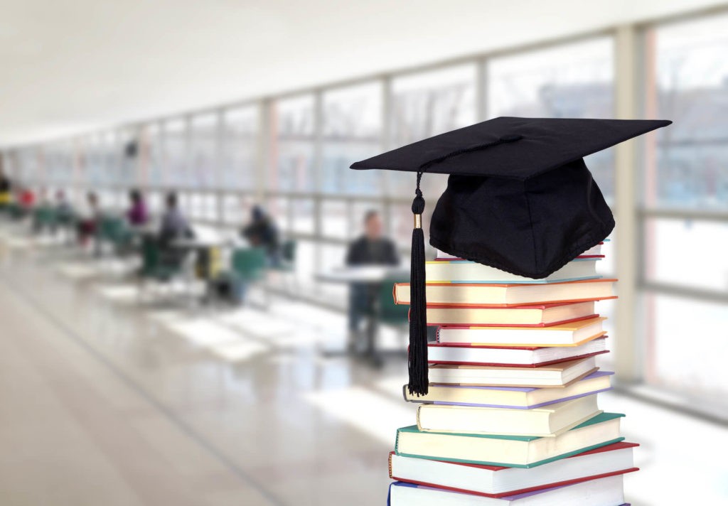 Graduation cap on top of a stack of books in a sunny hallway.