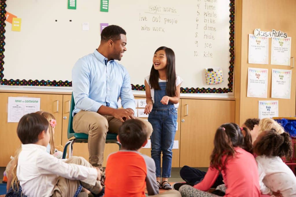 Teacher sitting with students with a young, smiling girl speaking to the group.