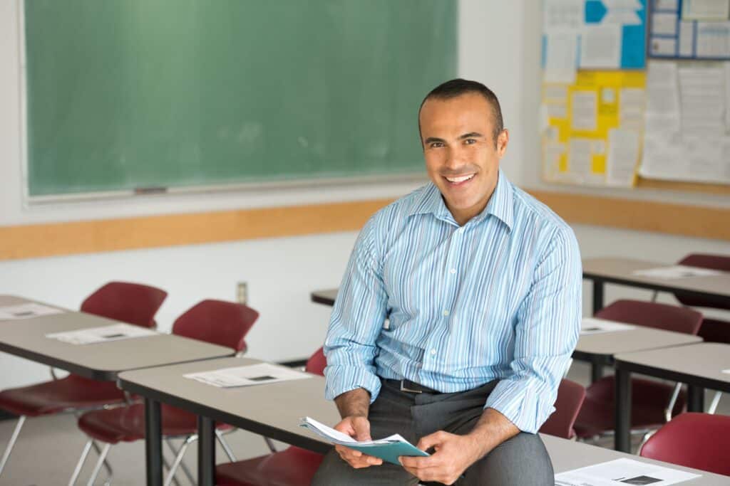A teacher sits on a desk in his classroom, smiling and holding a stack of papers.