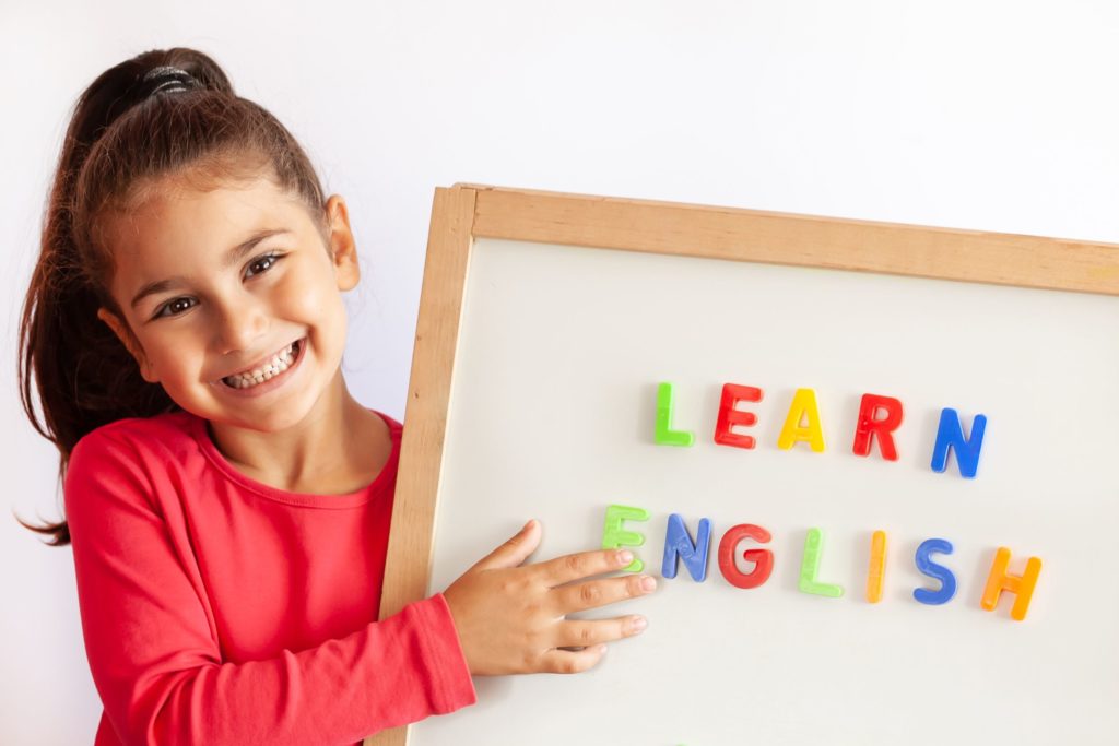 A young girl holds up a whiteboard with magnet letters spelling out “Learn English.”