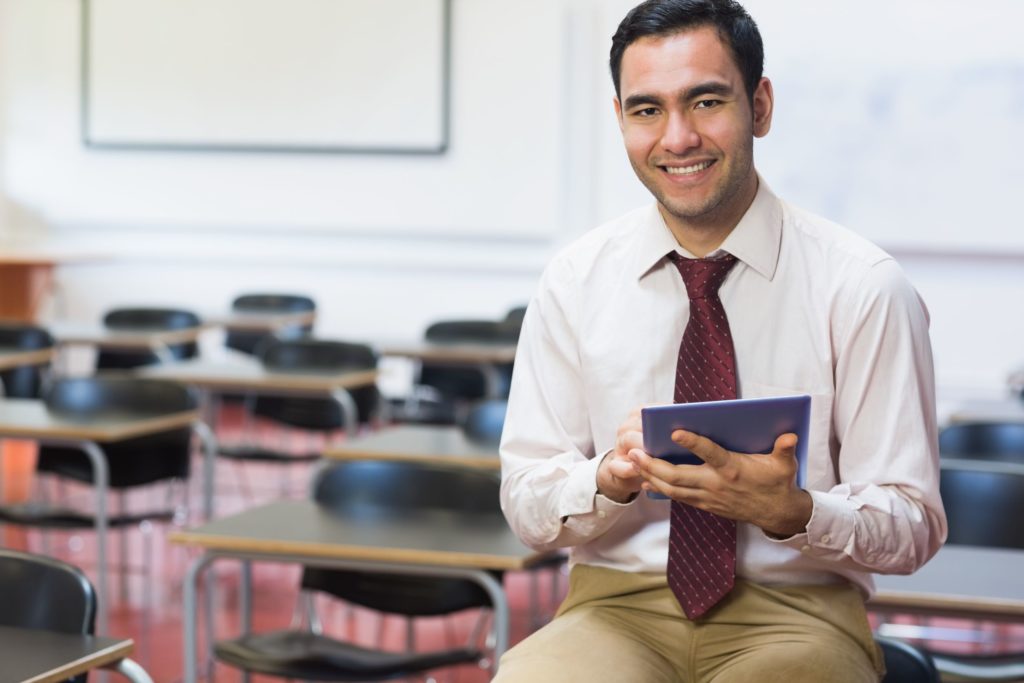 A young teacher sits smiling in his classroom on his tablet.