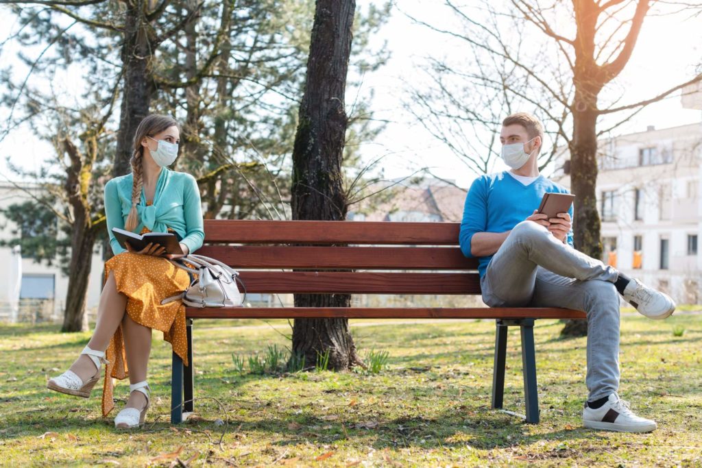 A man and woman social distance with masks on opposite sides of a college campus bench, discussing the books they have.