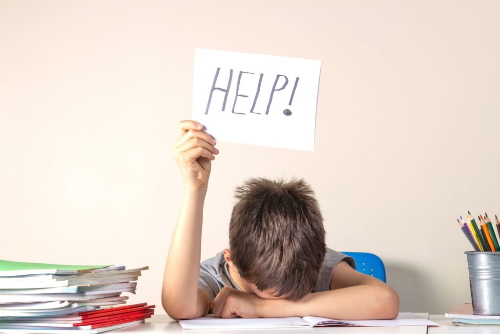 A boy sits at a school desk with his papers and books with his head down and holding up a sign that says “Help!”