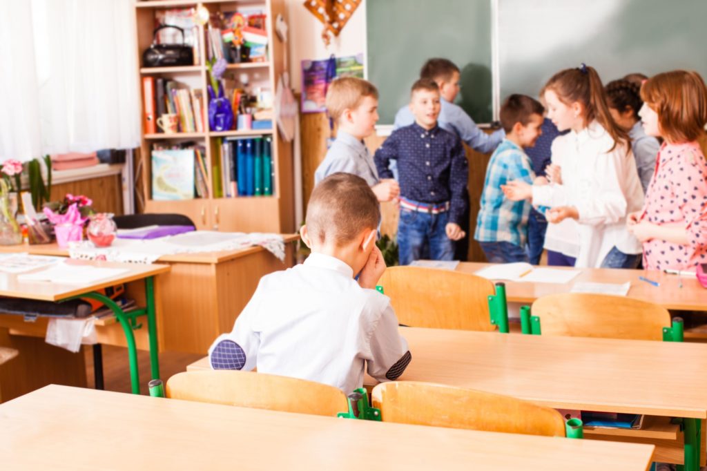 Young boy crying by himself at his desk while other children play.