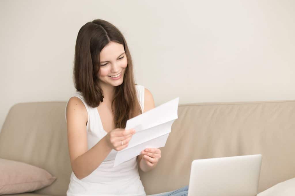 Young woman smiling and sitting on a couch opening a letter.