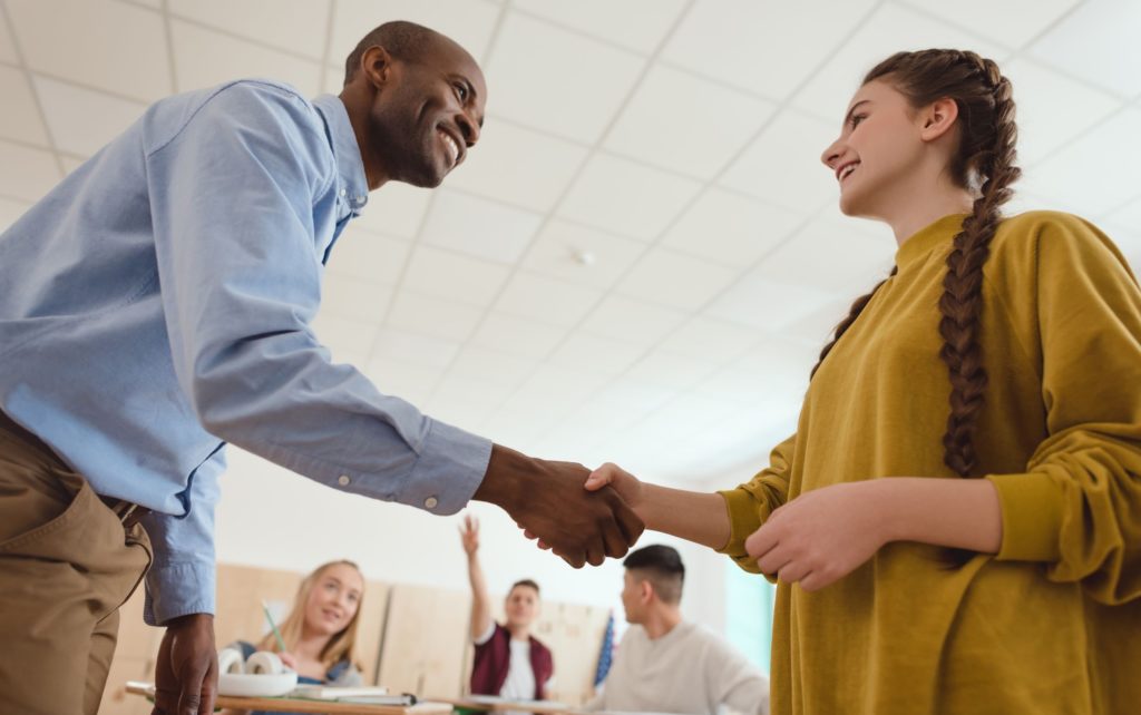 Male teacher shaking hands with a female student in a classroom.