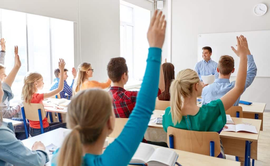 Classroom full of students with their hands raised while teacher sits up front.