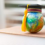 A graduation cap sits on top of a small globe, which is balanced on a textbook.