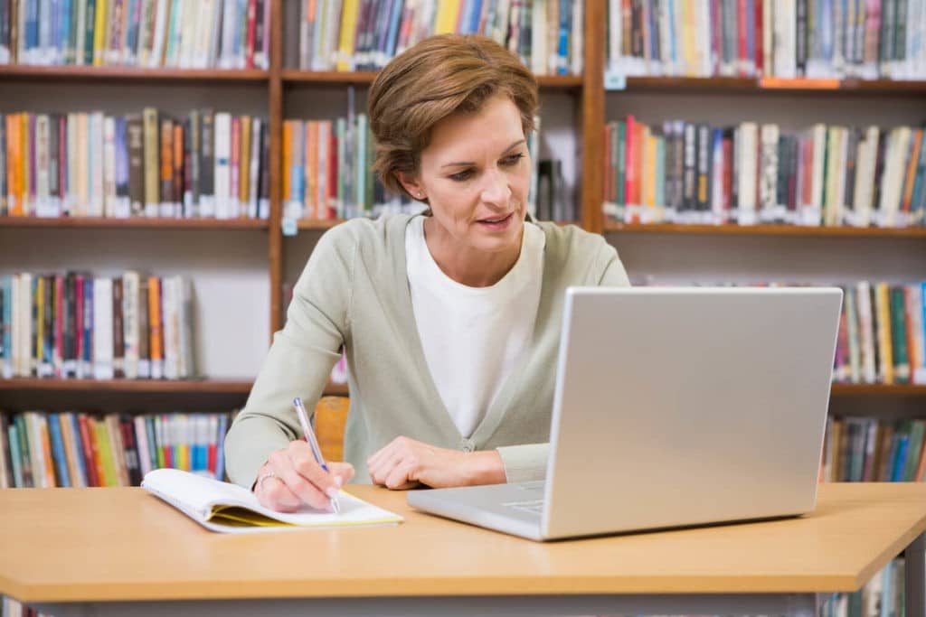 Teacher sitting in a library looking at a laptop while writing in a notebook.