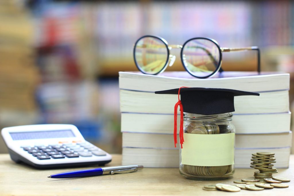 Jar of coins with graduation cap next to stack of books and calculator.