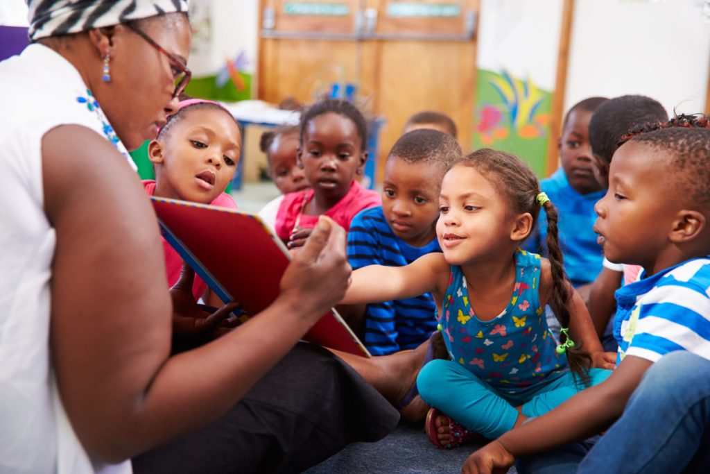A group of elementary students listen to their teachers read a book aloud.