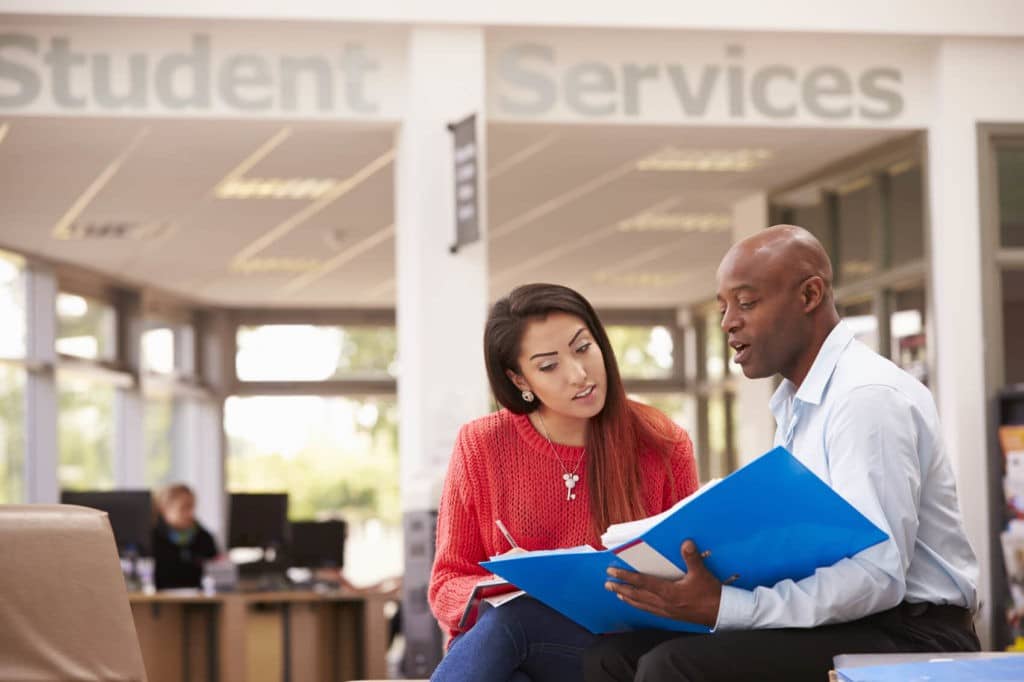 Man showing a binder to a woman, both sitting in front of student services.