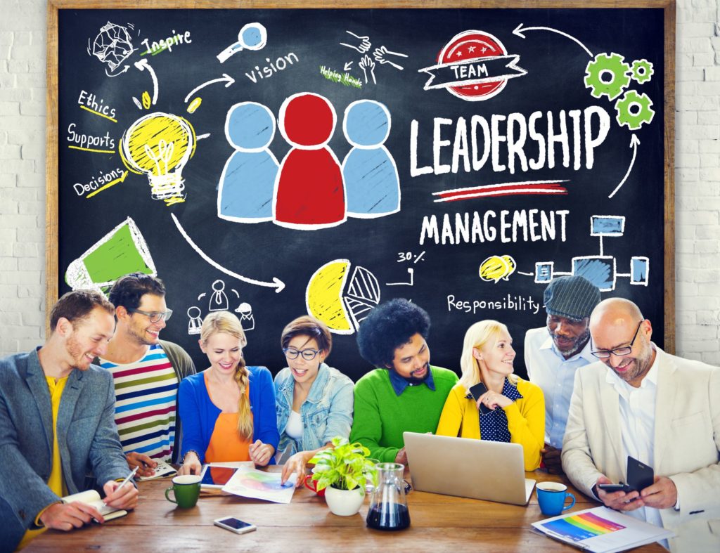 A group of professionals sit together in a meeting; the word “leadership” is drawn on a chalkboard behind them.