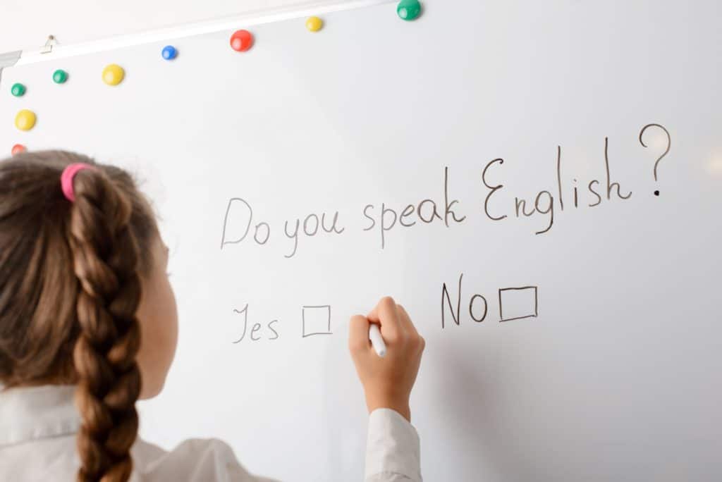 A young student looks at a whiteboard with the writing “Do you speak English? Yes or No” written on it.