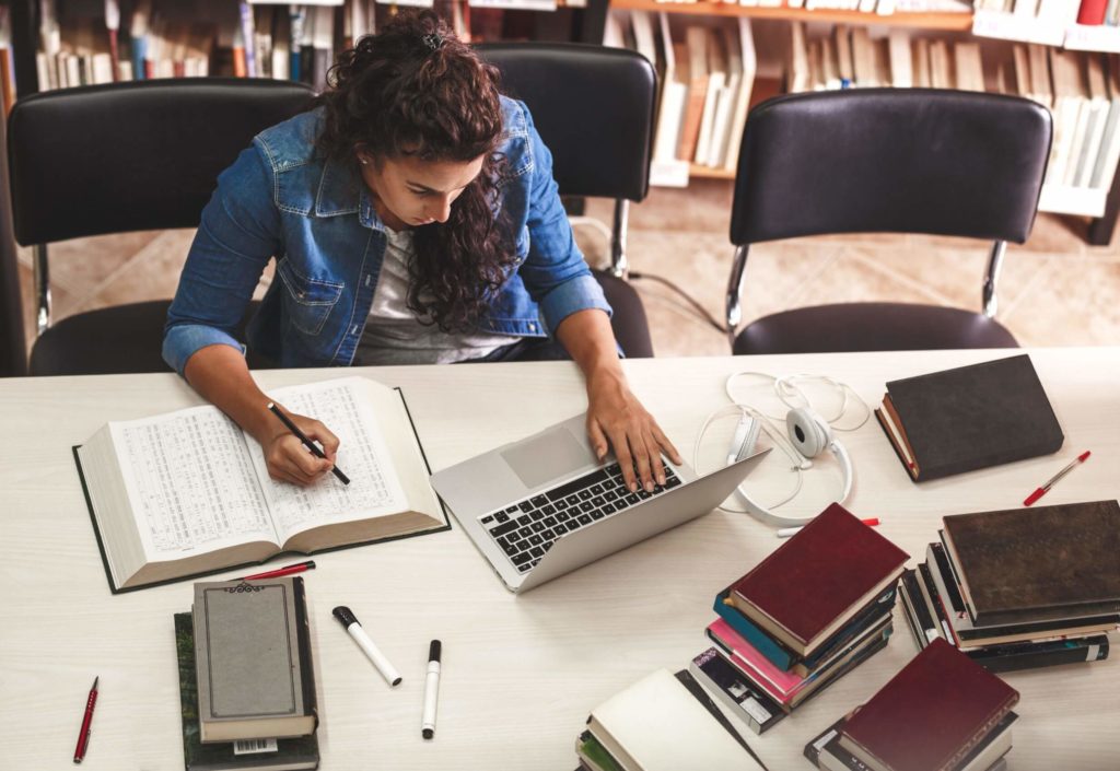 A female student sits surrounded by books and her laptop, doing research.