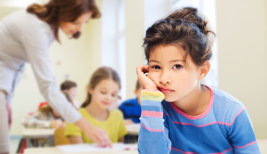 Young student looking bored in a classroom while a teacher helps other students.