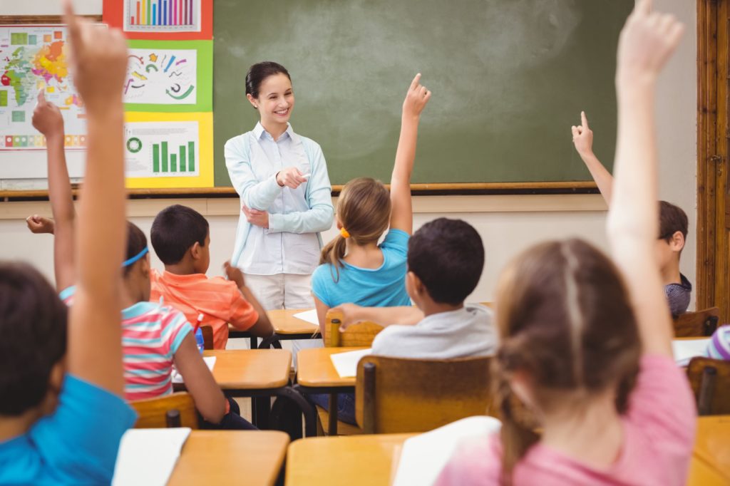 Smiling teacher at the front of a classroom full of students with their hands raised.