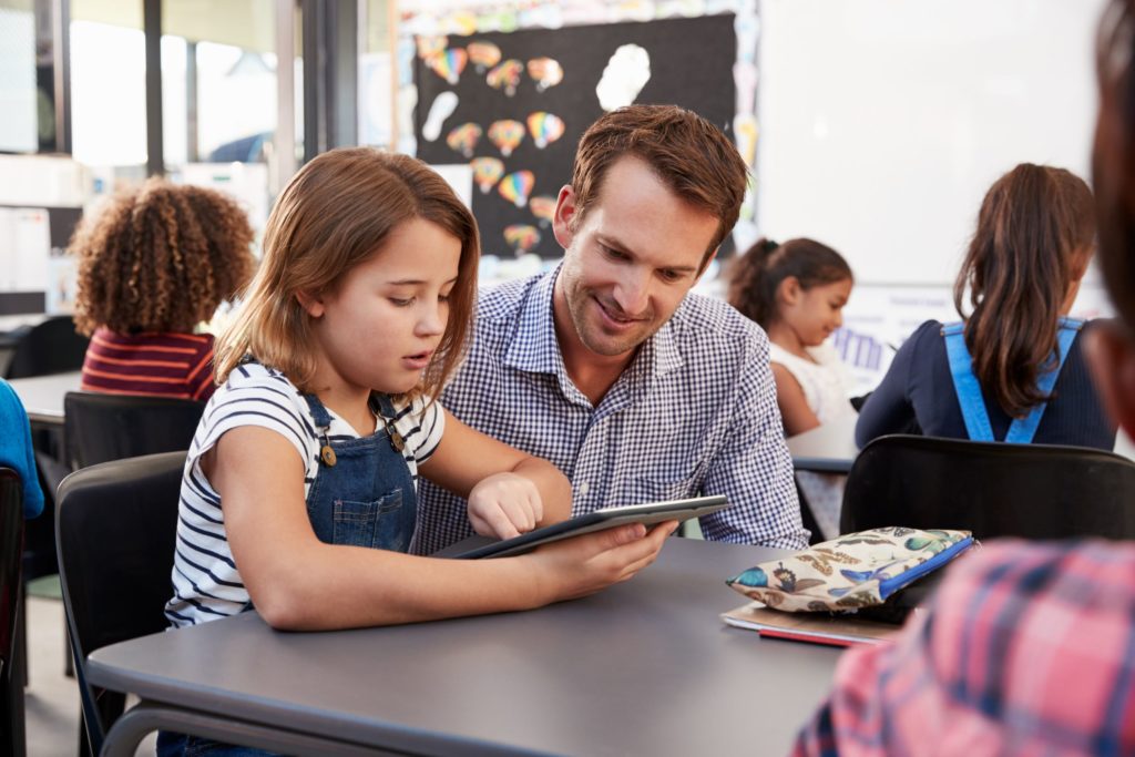 Young student sitting at a desk using a tablet with a male teacher kneeling next to her.