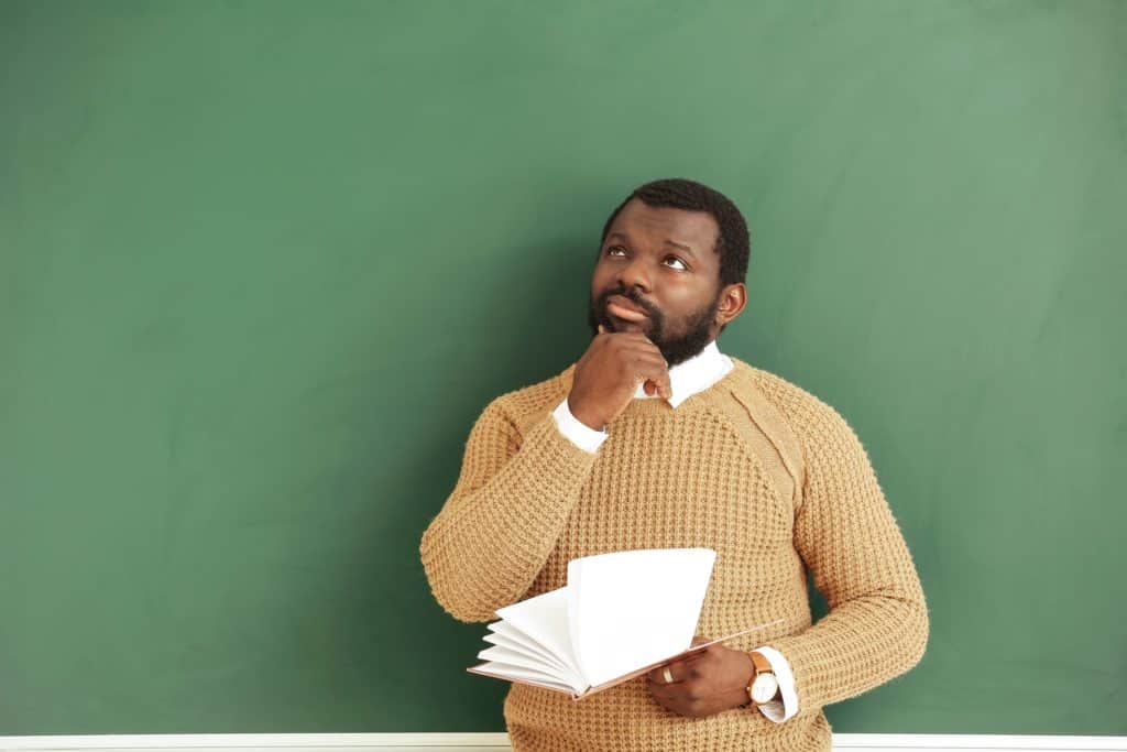 A teacher stands in front of a chalkboard, reflecting thoughtfully.