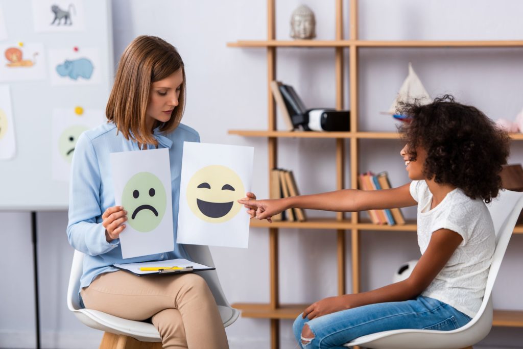 A girl is shown two faces on pieces of paper; she points at the smiling one to report her current emotion.