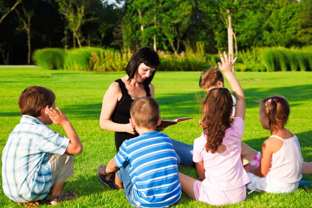 Group of young students sitting in the grass with a teacher reading a book to them.