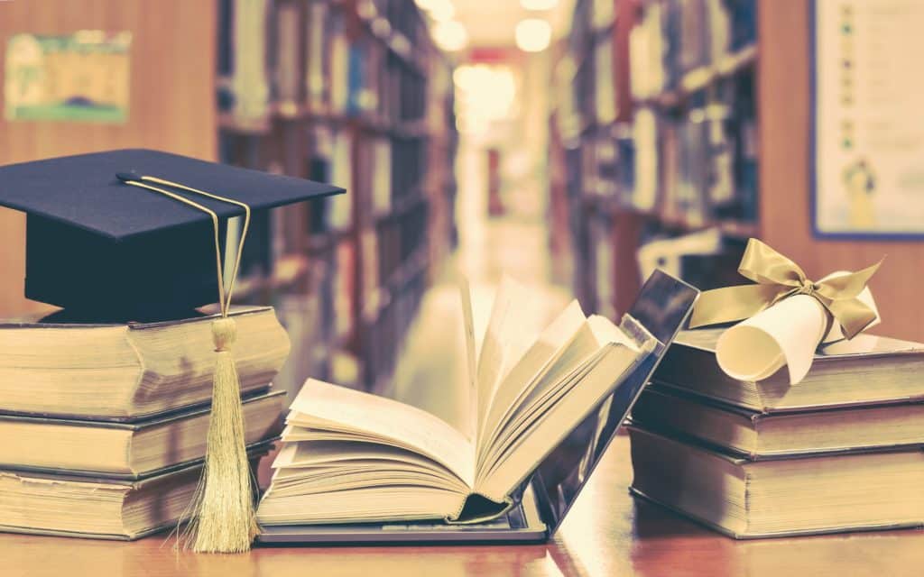 A stack of books and a laptop sit on a library desk between a graduation cap and diploma.