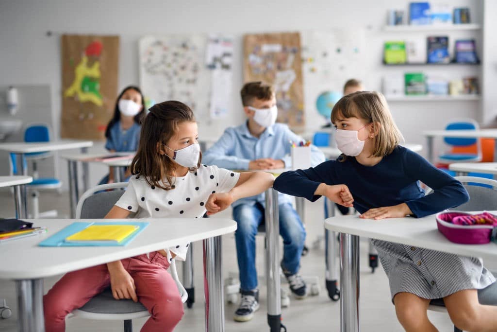 Two young girls in a classroom bumping elbows together.