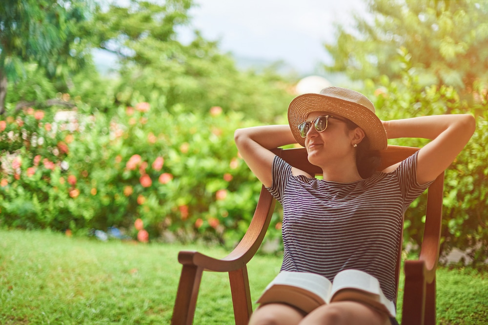 Women wearing a hat and sunglasses relaxing outside with a book.