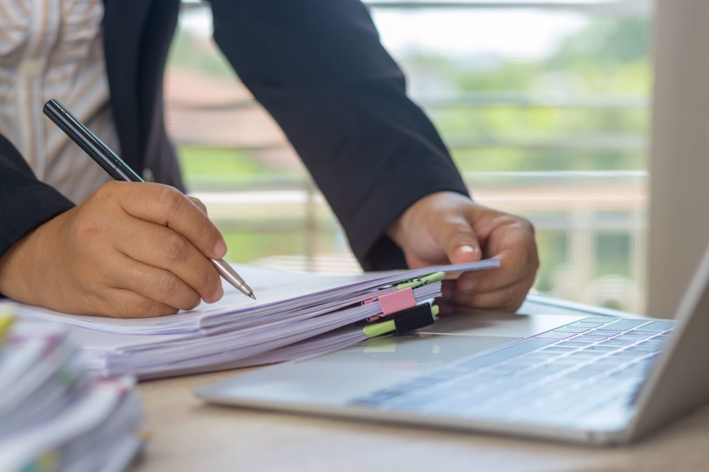 Adult writing on a stack of papers next to an open laptop.