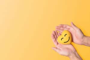 A pair of hands holds a yellow heart shaped stress ball that has a smiling face.