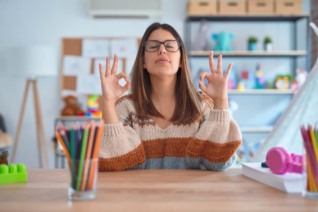 Young teacher sitting at her desk and meditating in a colorful classroom.