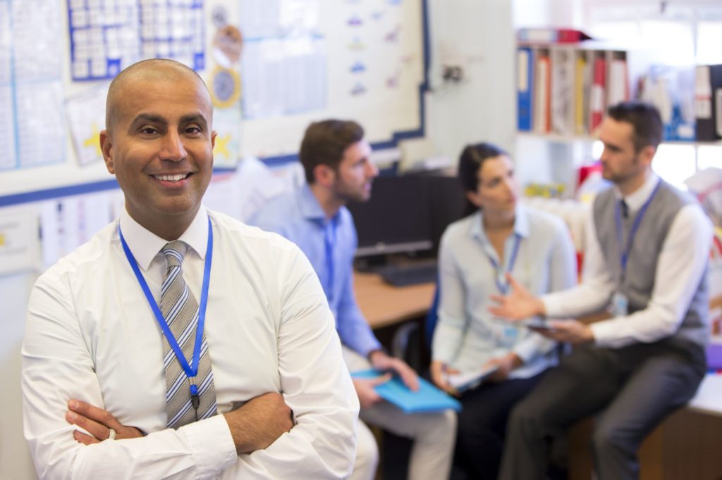 Male principal smiling with arms crossed in front of a group of teachers talking.