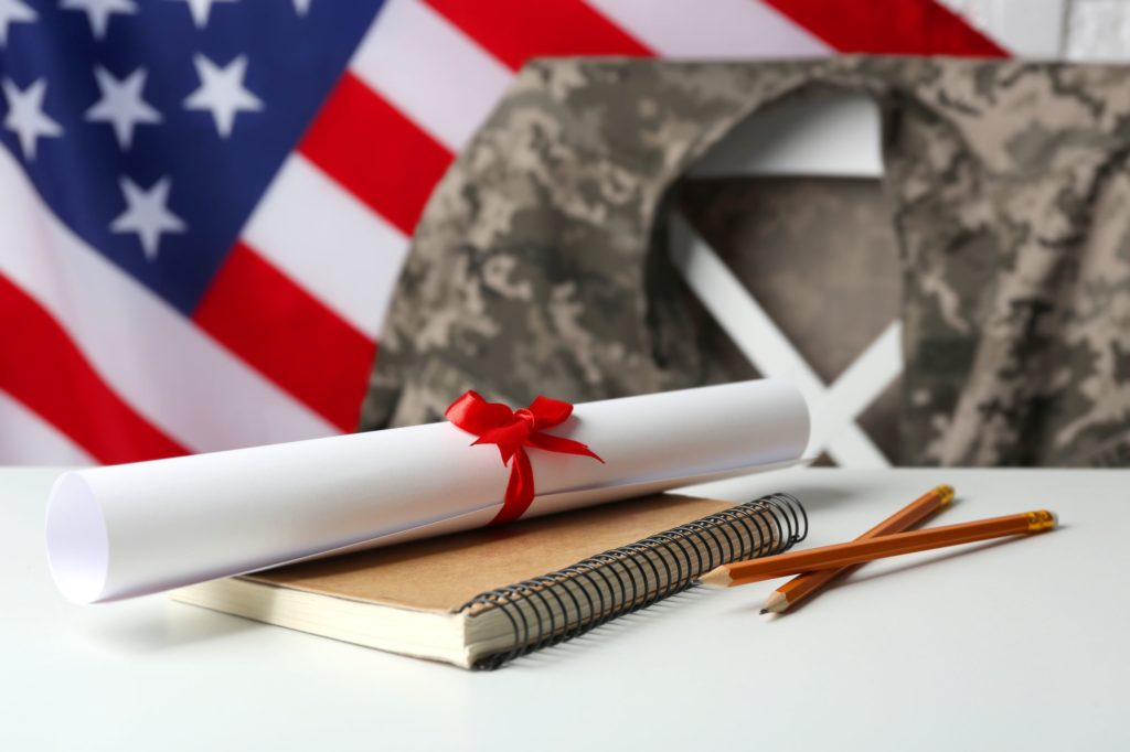 Army jacket on a chair in front of an American flag with a notebook, pencils, and diploma on a table.