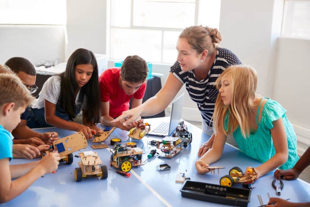 Students in an after-school program gather around a table as a teacher instructs them how to build a robot vehicle.