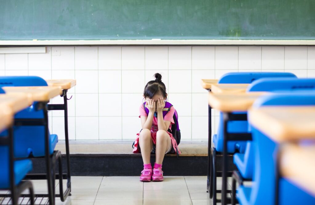 A young student sits at the front of an empty classroom, holding her head in her hands.