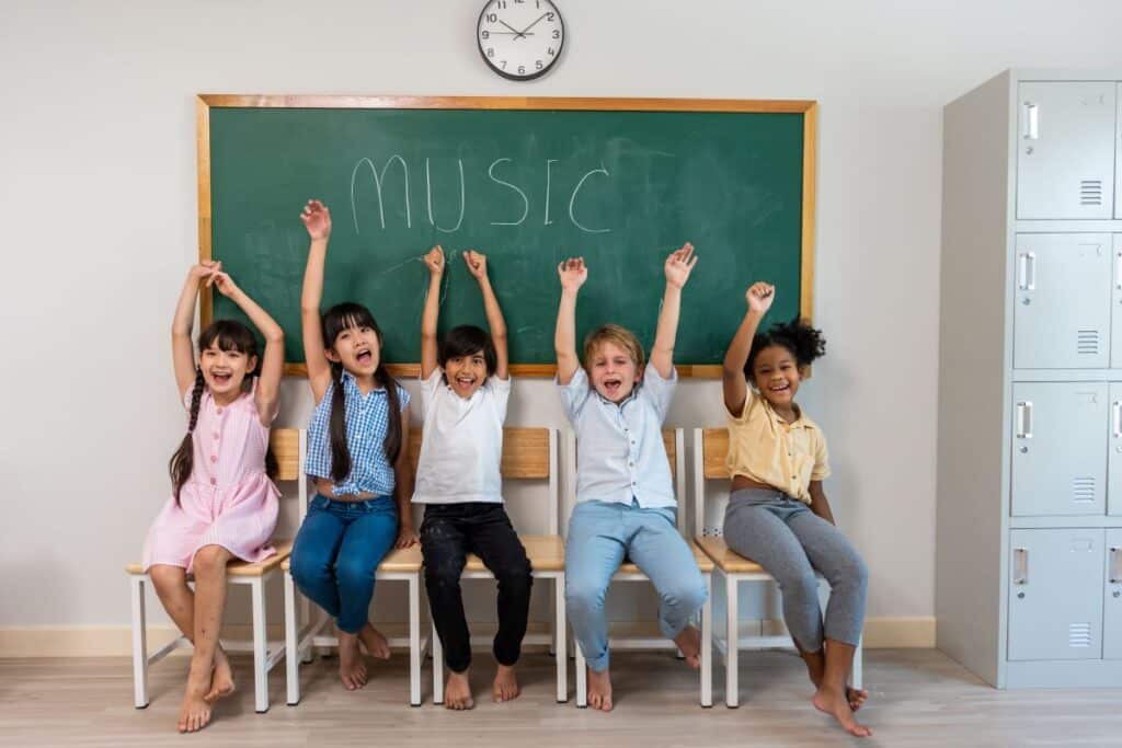 Student mental health: a group of students sit on a classroom table smiling and putting their hands in the air, the chalkboard has “music” written on it behind them.