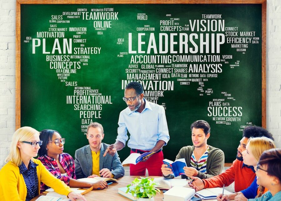 Man standing in front of a blackboard talking to colleagues seated at a table.