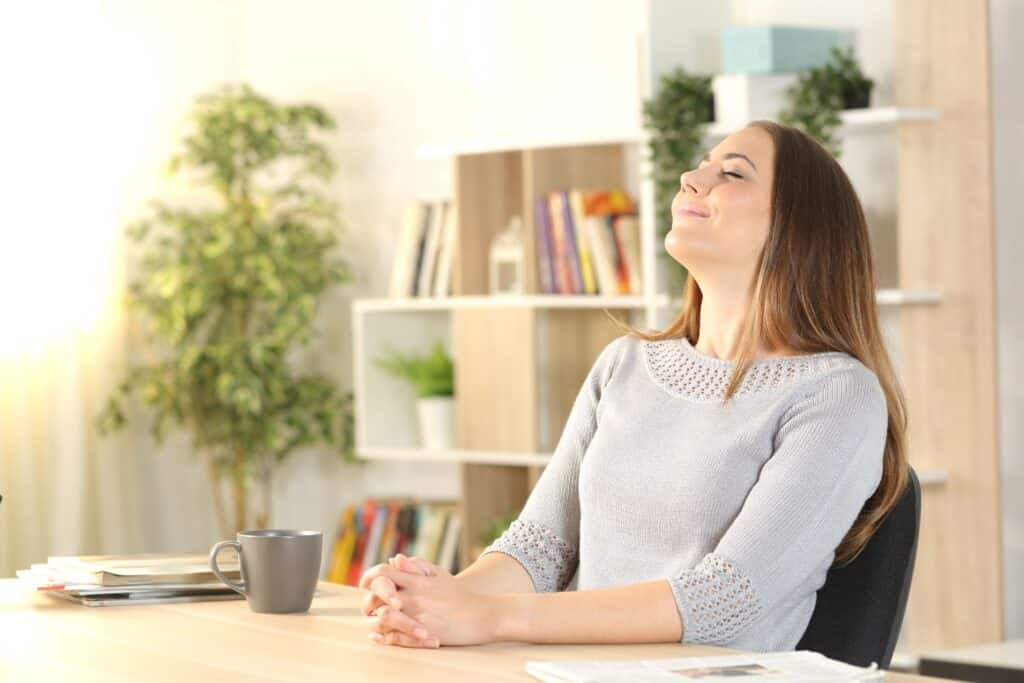 A relaxed woman sits at her desk, taking a deep breath and smiling.