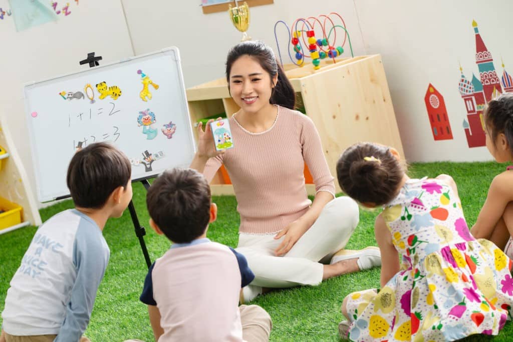 A kindergarten teacher sits on the floor teaching children in a classroom.