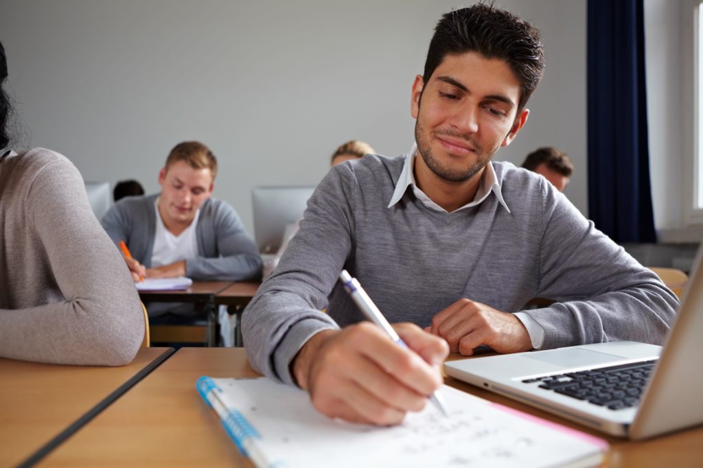 Young man taking notes in a notebook while using a laptop.