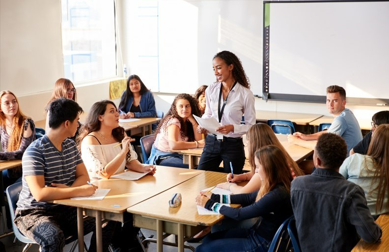 A female teacher smiles and instructs a diverse group of teen students.