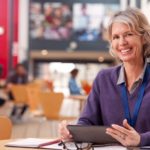 A teacher sits at a table, working on a tablet.